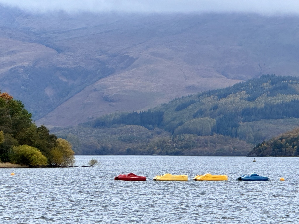 colourful paddle boats moored on loch, with distant hills and low cloud.