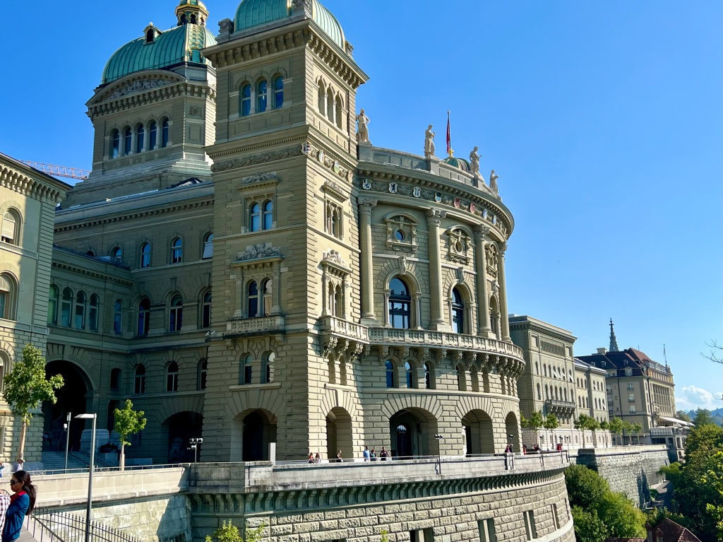 The ornate and rounded facade of the  Swiss parliament building, sitting atop a  high brick wall overlooking the river valley.