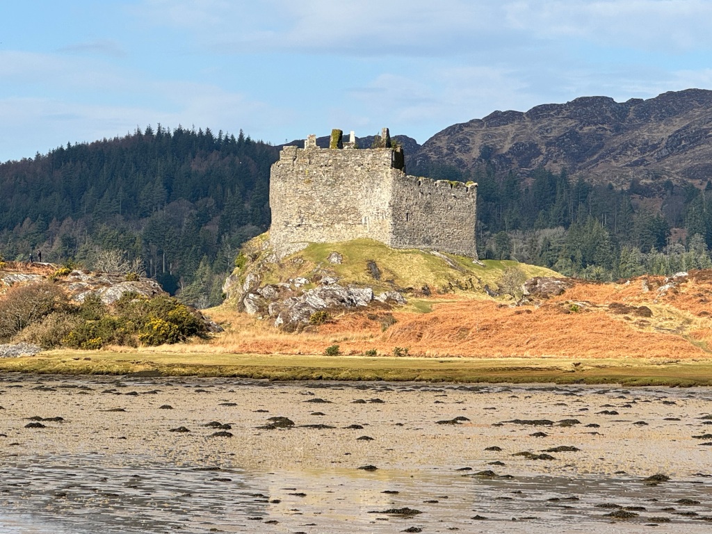 Tioram Castle ruins, set on island with sandy beach in foreground, and tree-covered hills behind