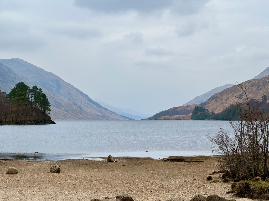View of Loch Shiel, with steep hills either side, a gravel beach in the foreground and grey sky.